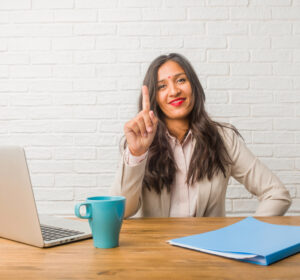 Woman holding up one finger at a computer indicating number 1. 